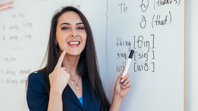 Woman pointing to her mouth while teaching pronunciation in front of a whiteboard with phonetic spellings written on it.