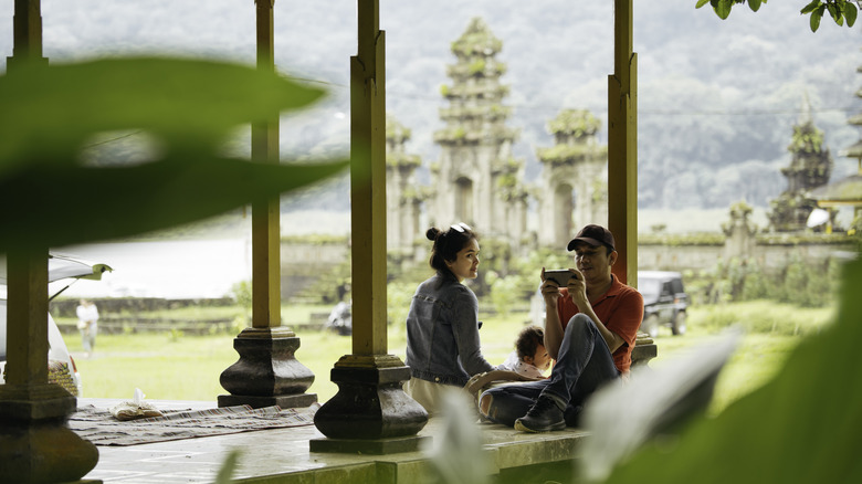 Family sitting under an open pavilion near a lakeside temple, with one person using a smartphone and mountains in the background.