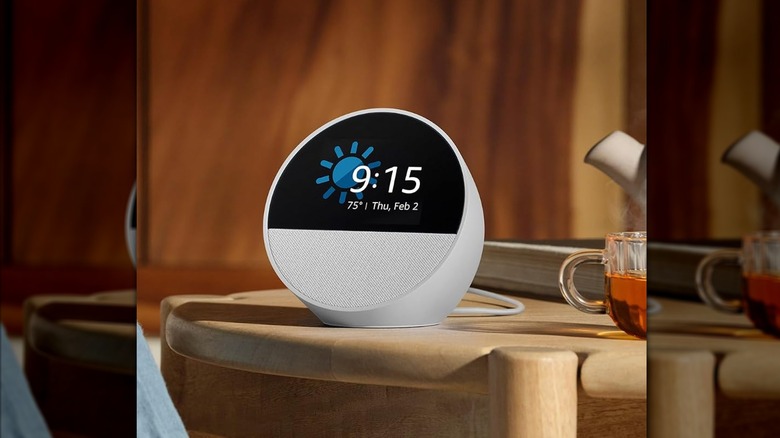 A white Amazon Echo Spot on a wooden side table next to a transparent cup of tea.