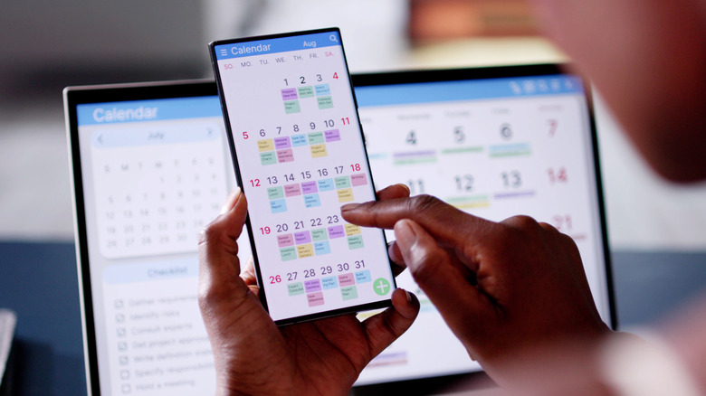 An over-the-shoulder view of a person accessing a digital calendar on their phone. Another digital calendar is visible on a larger screen in the background.