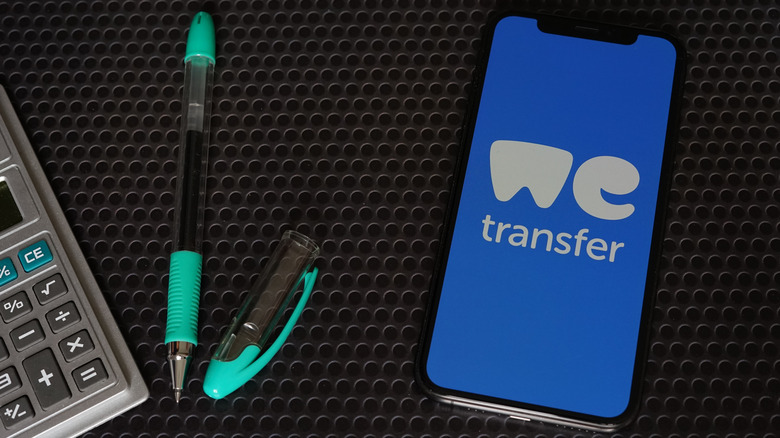 The WeTransfer logo open on a phone, next to two pens and a calculator on a dark surface