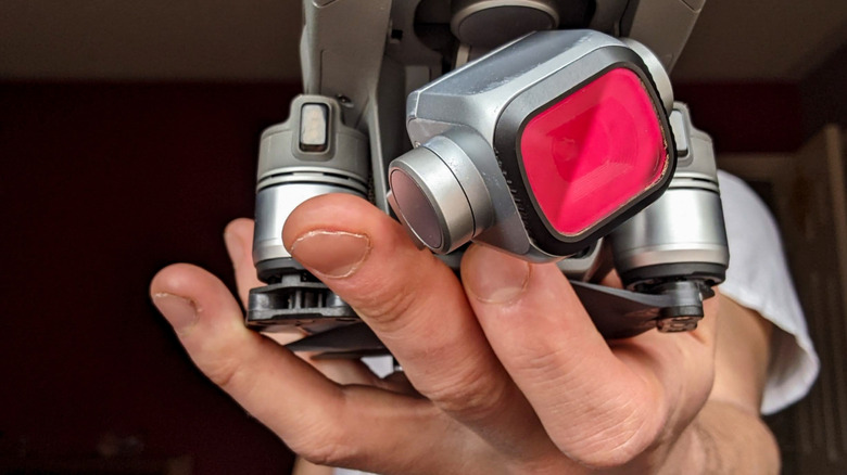 A close-up image of a compact camera drone being held in someone's hand, with a red-tinted camera filter attached to the gimbal-mounted lens.