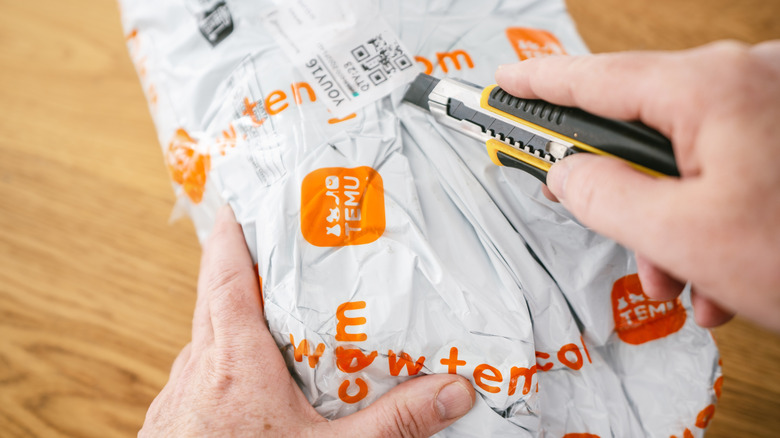 Person using a utility knife to open a Temu shipping package, with the plastic mailer held on a wooden table.