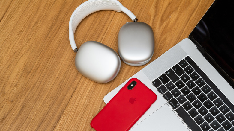 Apple AirPods Max headphones resting beside a MacBook and an iPhone in a red case on a wooden desk, illustrating an Apple ecosystem workspace.
