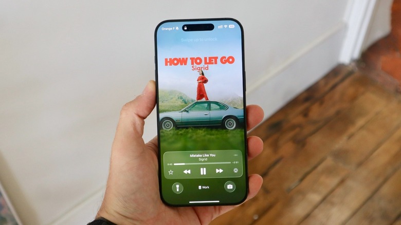 A hand holding an iPhone 16 Pro Max. On the display, iOS 26's animated cover Lock Screen