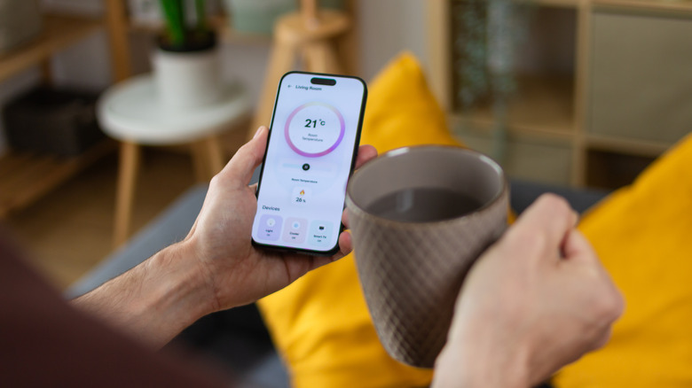 A person checking the smart thermostat app on their phone while holding a coffee mug