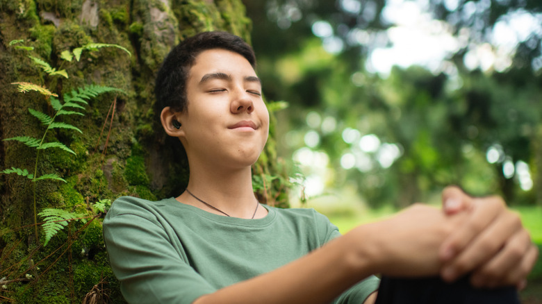 A boy wearing earbuds relaxing next to a tree.