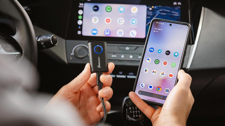 A person holding an AAWireless dongle next to a smartphone with a card dashboard in the background.