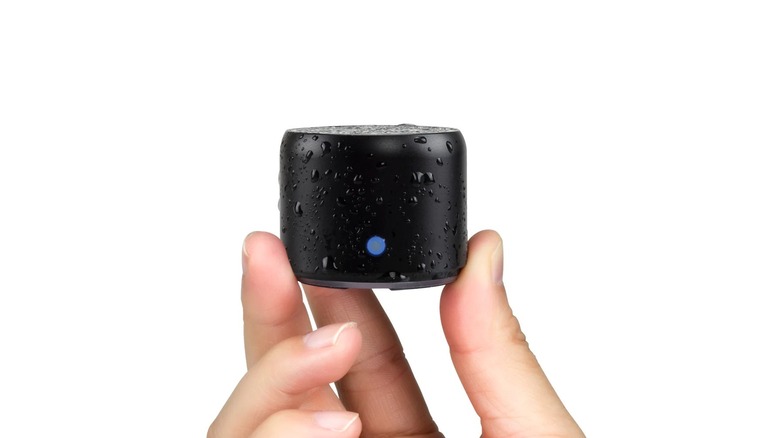 A small, cylindrical black speaker with water droplets on its surface is held between two fingers, with a blue indicator light visible on the front against a plain background.