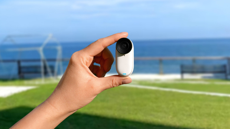 A small, pill-shaped action camera is held between fingers in the foreground, with a grassy area and ocean view blurred in the background.