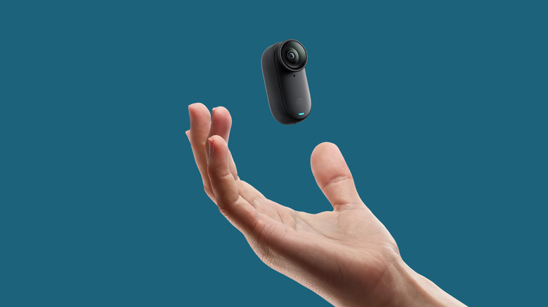 A small, compact action camera appears suspended above an outstretched hand against a solid blue background.