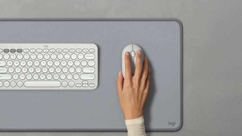 A minimalist desk setup shows a compact white keyboard and a matching mouse on a large gray desk mat, with a hand resting on the mouse.