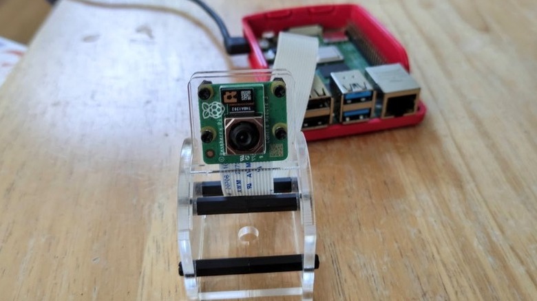 A close-up image of a Raspberry Pi 5 camera module connected to a Raspberry Pi 5 sitting in a case in the background.