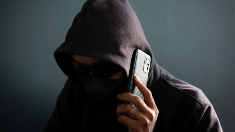 Person in a dark hoodie and face covering holding a smartphone to their ear, with their face mostly obscured in shadow.