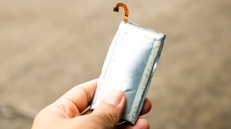 Hand holding a swollen lithium battery pack with a partially exposed connector, showing visible bulging and damage.