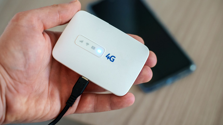 Hand holding a small portable 4G mobile hotspot device connected by USB, with a smartphone blurred in the background.
