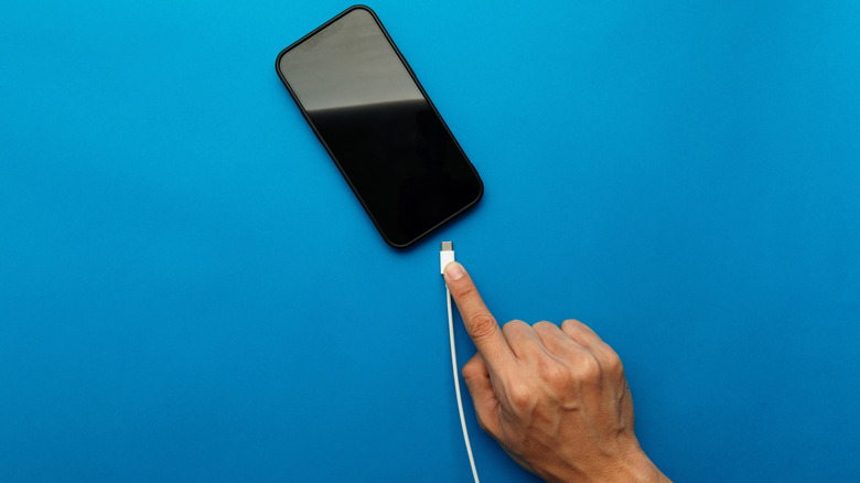 Hand holding a charging cable and about to plug it into a smartphone lying on a blue surface.