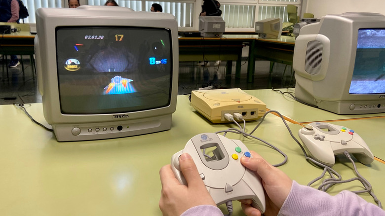 Person playing a Sega Dreamcast on a CRT monitor, holding the original controller while a racing game runs on screen, with another setup visible in the background.