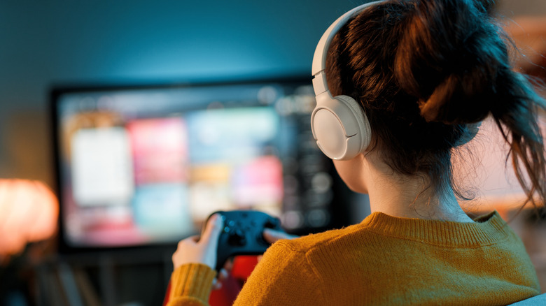 Rear view of a person wearing over-ear headphones and holding a game controller, seated in front of a TV displaying a blurred interface.