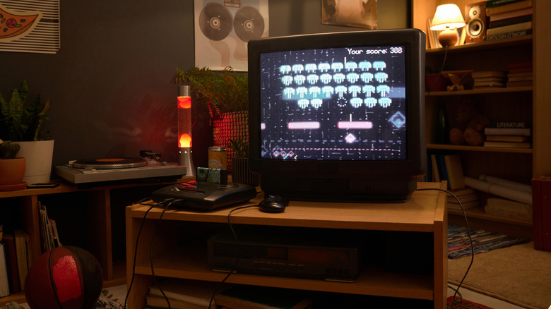 Cozy retro living room with a CRT TV showing a Space Invaders-style game, a game console on a wooden stand, a glowing lava lamp, turntable, plants, and bookshelves in the background.