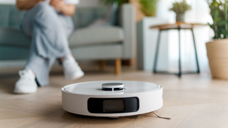 A white robot vacuum cleans the floor while a person sits on a couch behind it