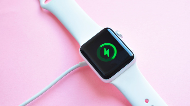 A white banded Apple Watch displays a green lightning bolt "charging" icon in front of a blush pink background.