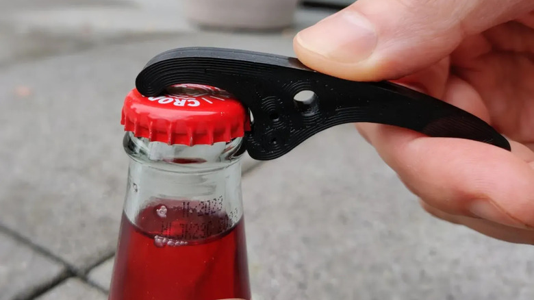 A person holding a black 3D-printed bottle opener against the cap of a glass bottle