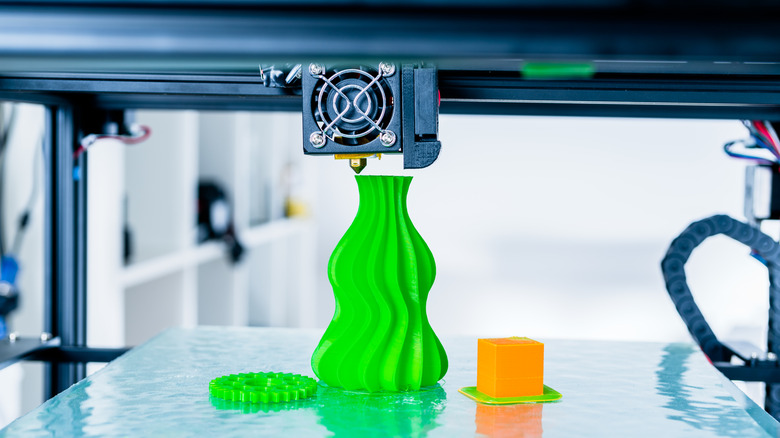 A lime green 3D-printed vase sits on a 3D printer bed beside a green gear and orange cube
