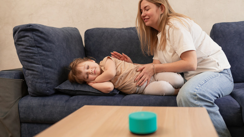 An Alexa speaker on a table in front of a child sleeping on the couch with an adult sitting next to them