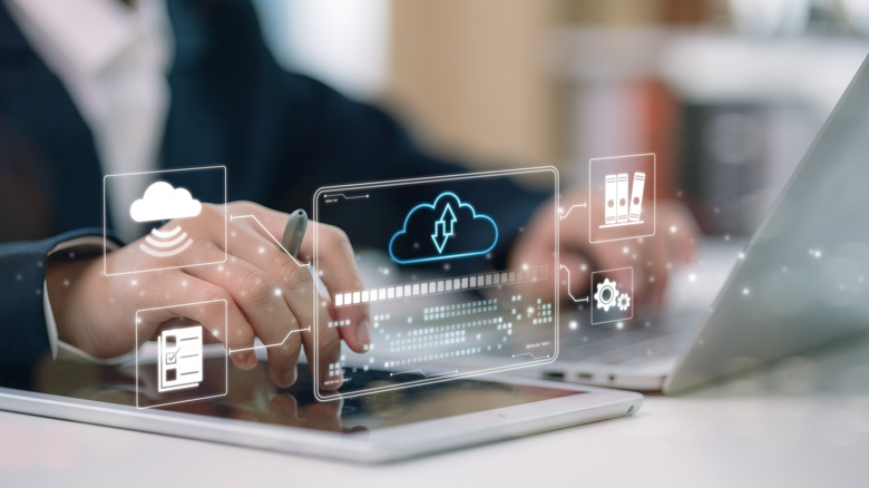 A person interacting with a laptop and a tablet, with a holographic overlay showing icons for cloud computing