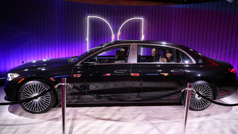 A black luxury sedan with four people inside the car parked indoors in front of a large LED Dolby logo.