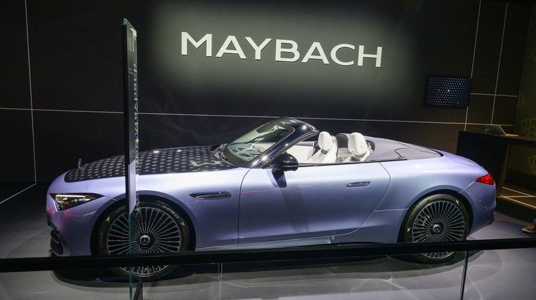 A metallic periwinkle Maybach convertible with white seats in front of a glowing Maybach logo.