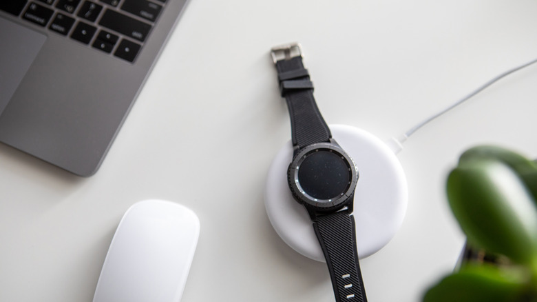 A smartwatch on a wireless charger next to a laptop computer, all on a white surface.