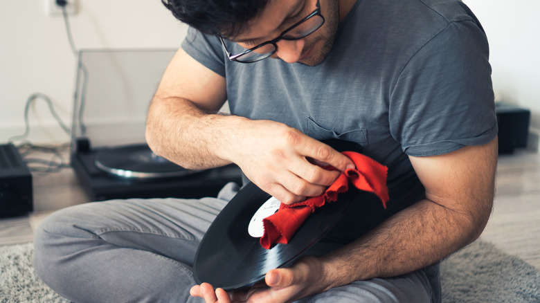 Person cleaning a vinyl record with a microfiber cloth while sitting on the floor, with a turntable visible in the background.