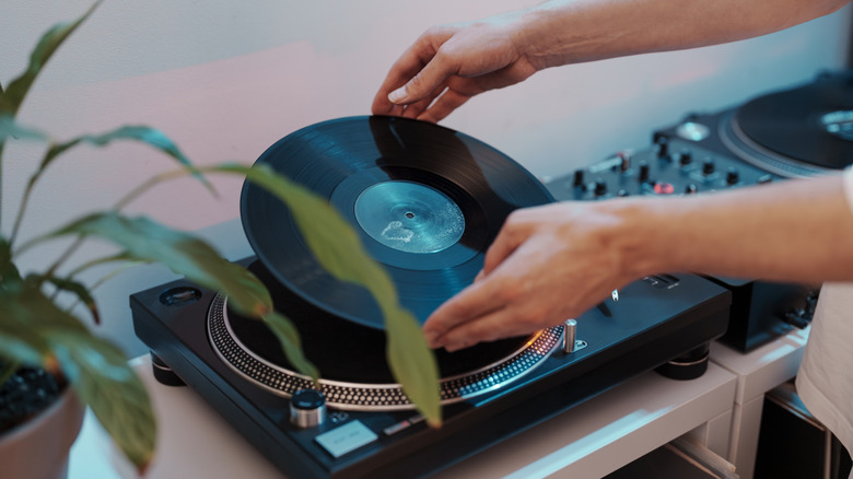 Person placing a vinyl record onto a turntable, with a DJ-style audio setup visible in the background and a houseplant partially framing the scene.