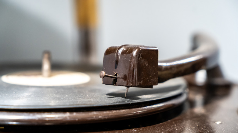 Close-up of a turntable tonearm and crude stylus resting on a vinyl record, with the needle positioned in the record groove and the background softly out of focus.