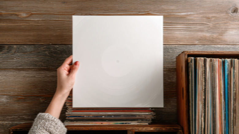 Hand holding a blank vinyl record sleeve above a stack of records, with a crate of vinyl albums and a wooden tabletop visible in the background.