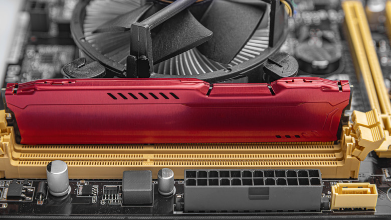 Close-up of a red desktop RAM module installed in a motherboard DIMM slot, with a CPU air cooler and surrounding PC components visible in the background.