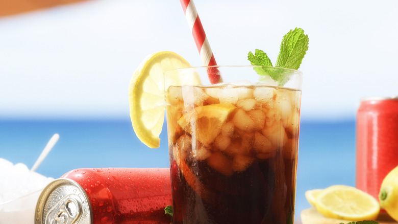 A glass of soda with ice and lemons, surrounded by cold soda cans with the ocean in the background.