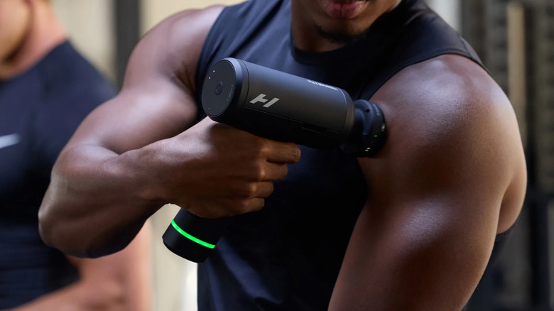 A muscular man applies a massage gun to his arm.