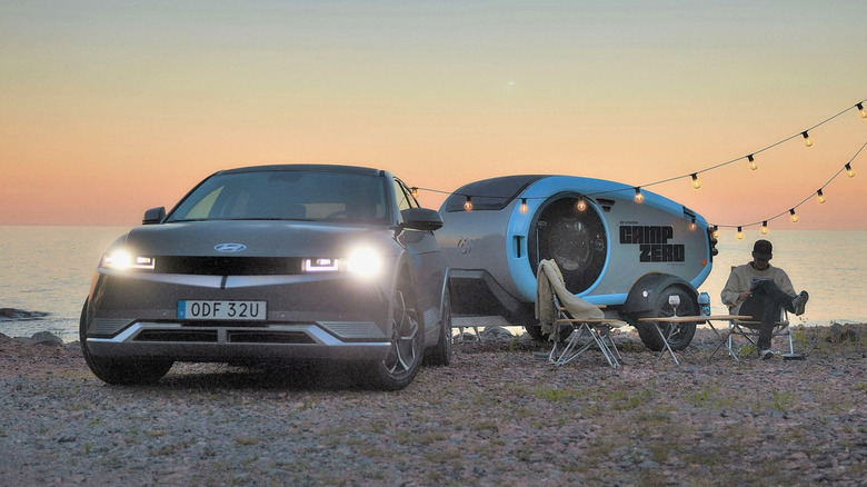 The Ioniq 5 with its headlights on as it charges string light bulbs beside a small camping trailer by a lake.