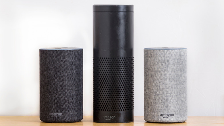 Three Amazon Echo speakers beside each other on a wooden surface in front of a white wall.