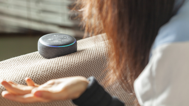 A person with long brown hair leaning over, extending an open palm to the side, and looking at an Amazon Echo Dot speaker.