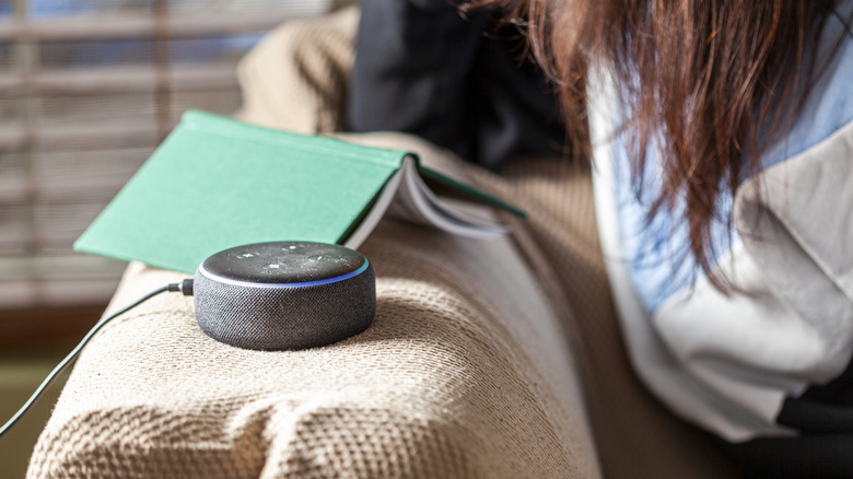 An Amazon Echo Dot speaker on top of a beige couch next to a green hardcover book and a person seated on the couch.
