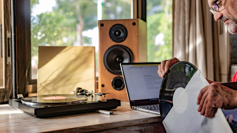 A man holding a vinyl record near a turntable, laptop, and large loudspeaker in the background.