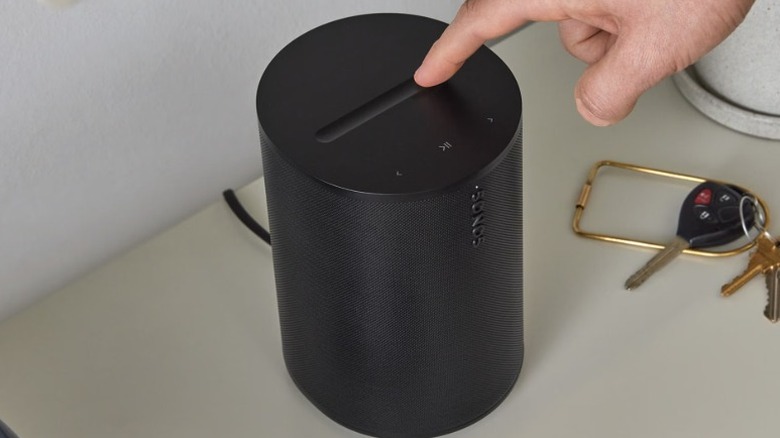 A person interacting with the Sonos Era 100 SL which is standing on a table. A pair of keys sit beside the speaker.