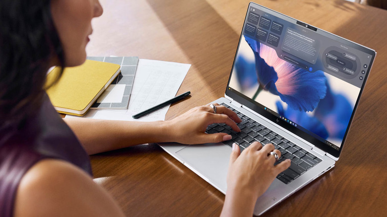 A person types on a slim laptop at a wooden desk, with notebooks and a pen placed nearby.