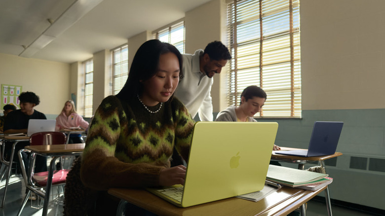 A student sits at a classroom desk using a bright yellow laptop, focused on her work.