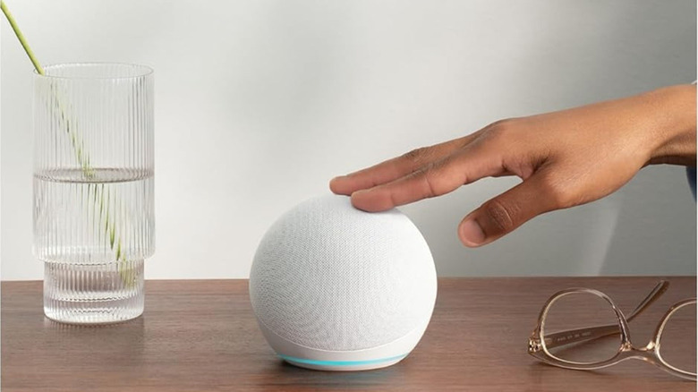 A person pressing the top button of the Amazon Echo Dot (5th Gen) smart speaker, which is on a wooden table beside a glass vase and eyeglasses.