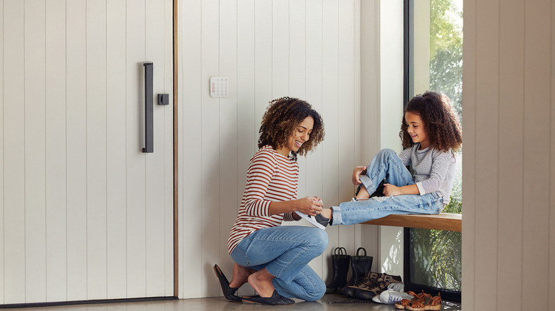 A mother tying the shoes of her daughter, with the Ring Alarm Keypad shown outside their door.